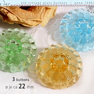 Può includere: Tre bottoni rotondi in vetro di colore verde chiaro, azzurro e giallo. Ogni bottone presenta un motivo a punti in rilievo e un bordo smerlato. Il testo 3 buttons e ø je ca 22 mm è stampato sotto i bottoni, con altro testo sopra.