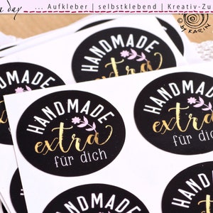 Pode incluir: Uma pilha de adesivos redondos pretos e brancos com o texto "HANDMADE extra für dich" em letras douradas. Os adesivos têm um design floral rosa.