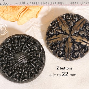 Könnte beinhalten: Zwei antike Glas Knöpfe, jeweils ca. 22 mm Durchmesser, mit detaillierten Mustern. Einer ist dunkelgrau mit einem Spiralmuster, der andere eine Mischung aus Gold und Dunkelgrau mit einem floralen Motiv. Der Text lautet "2 Knöpfe".