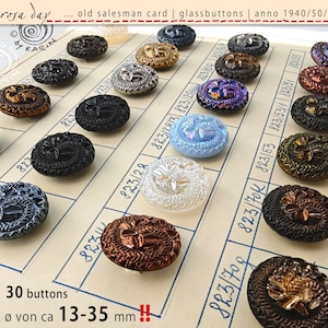 May include: A selection of 30 vintage glass buttons in various colours, including black, brown, gold, and iridescent. The oval-shaped buttons feature intricate floral designs and measure approximately 1.3-3.5 cm in diameter. The buttons are displayed on an old salesman card.