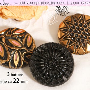 Puede incluir: Tres botones redondos de vidrio vintage con un diámetro de 22 mm. Un botón presenta un patrón negro y dorado con detalles en rosa, otro es negro sólido con un diseño radial de rayos de sol y otro es dorado con un patrón de estrella central.