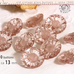 Op de afbeelding: Een verzameling van tien vintage glazen knopen in een lichtroze tint. Elke knoop heeft een ronde vorm met een getextureerde, gefacetteerde rand en een centraal verhoogd patroon dat op een bloem lijkt. De tekst 10 buttons en ø je ca 13 mm is zichtbaar.