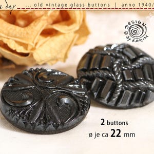 Puede incluir: Dos botones redondos de vidrio negro con complejos patrones de relieve moldeados. Un botón presenta un diseño de volutas en espiral, mientras que el otro muestra un patrón geométrico de barras. El texto indica 2 botones de 22 mm de diámetro.