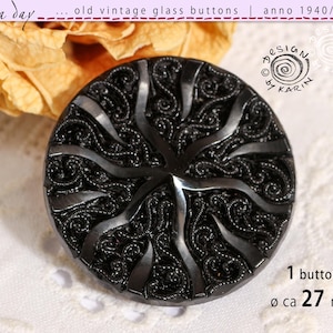 May include: A black, vintage glass button with an intricate, swirling design. The button is approximately 2.7 cm in diameter and features the text "DESIGN by KARIN" and "1 button". The text "old vintage glass buttons anno 1940/50/60" is also visible.