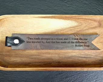 May include: A brown leather bookmark with a black leather strap and a metal button. The bookmark features an engraved quote from Robert Frost's poem "The Road Not Taken": "Two roads diverged in a wood, and I - I took the one less traveled by, And that has made all the difference."