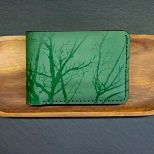 May include: A green leather wallet featuring a tree silhouette design. The wallet is stitched with dark thread and has a smooth, textured surface.