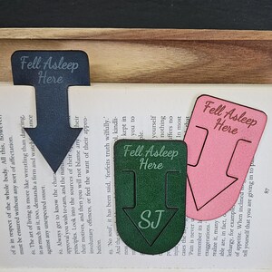 May include: Three wooden bookmarks with the text "Fell Asleep Here" in different colours: black, pink and green. The green bookmark has the initials "SJ" in white.
