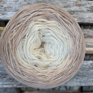 Peut inclure: Une pelote de laine dans un dégradé de beige, crème et blanc. La laine est enroulée en forme de boule et est prête pour le tricot ou le crochet.