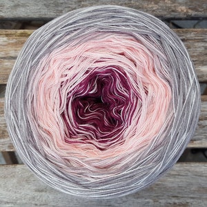 Op de afbeelding: Een bol garen met een kleurverloop, beginnend met lichtgrijs, overgaand naar lichtroze, dan donkerroze en eindigend met een diepe bordeaux.