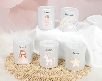 May include: Five white ceramic piggy banks with personalized names: Fiona, Hannah, Emilia, Carletta, and Theresa. Each bank features a different design: a ballerina, a unicorn with a rainbow, a princess, a unicorn, and a star.