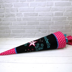 May include: A black and pink school cone with a blue star and the name "Isabella" embroidered on it. The cone is decorated with white stars and a blue ribbon.
