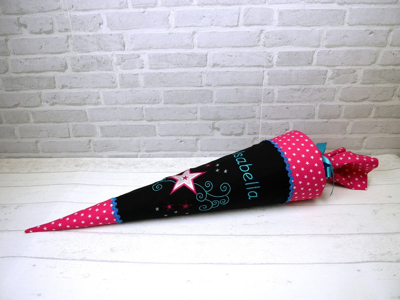 May include: A black and pink school cone with a blue star and the name "Isabella" embroidered on it. The cone is decorated with white stars and a blue ribbon.
