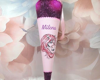 May include: A pink and purple school cone with a unicorn illustration and the name "Milena". The cone is decorated with a pink ribbon and a scalloped edge. The top is a dark purple fabric.