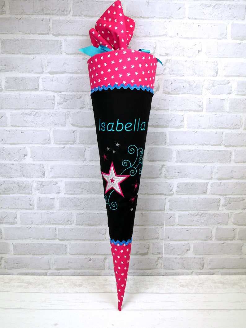 May include: A pink and black school cone with white stars and a blue ribbon. The cone has the name "Isabella" embroidered on it.