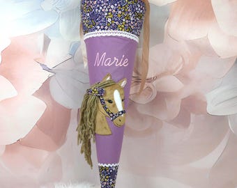May include: A purple cone-shaped school bag with a horse applique and the name "Marie" in script. The top and bottom are patterned with a floral design. The bag is tied with a ribbon.