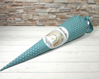 May include: A teal blue fabric cone with white stars, decorated with a white circle sticker featuring a rainbow, flowers, and the text "Mein 1. Schultag September 2023" and "Charlotte".