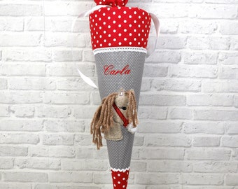 May include: A gray and red school cone with white polka dots. The cone has a white ribbon and a white lace trim. The cone is decorated with a brown and white horse and the name "Carla" is embroidered on the front.