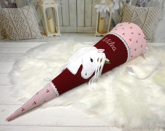 May include: A red and pink school cone with a white unicorn applique and the name "Milea" embroidered on the side. The cone is decorated with white lace and pink polka dots.