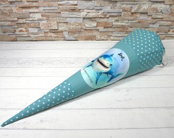 May include: A teal blue fabric cone with white stars and a shark graphic. The cone has a white label with the name "Erik" and the text "1. Schultag 2023".