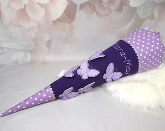 May include: A purple and white school cone with white polka dots and a silver trim. The cone has a purple butterfly design and the name "Clara-May" embroidered on the side.