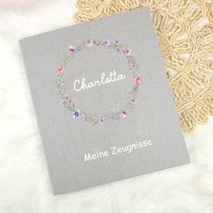Könnte beinhalten: Ein graues Notizbuch mit dem Namen "Charlotta" in weißer Schrift, umgeben von einem floralen Kranz. Der Text "Meine Zeugnisse" steht darunter. Das Notizbuch liegt auf einer weißen, flauschigen Oberfläche.