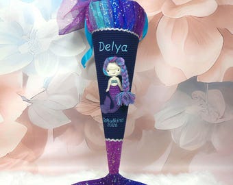 May include: A dark blue cone-shaped gift bag featuring a mermaid applique, the name Delya, and the text Schulkind 2026. The top is finished with a shimmering blue and purple tulle fabric, and the base is a purple and blue glittery mermaid tail.