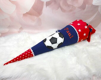May include: A blue, red, and white school cone with a soccer ball design. The cone is decorated with red stars and the name "Paul". The cone is placed on a white, fluffy surface.