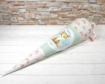 May include: A cone-shaped school bag with a fox illustration and the name "Flora". The bag features a light green and white polka dot pattern, and a pattern of rainbows and gold dots. A pink ribbon is attached to the top.