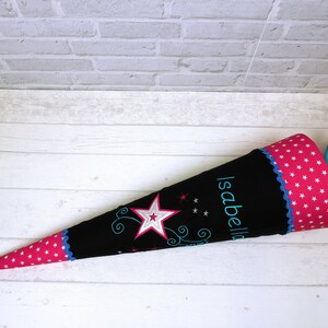 May include: A black and pink school cone with white stars and the name "Isabella" embroidered on it. The cone has a pink and white polka dot pattern and a blue ribbon.