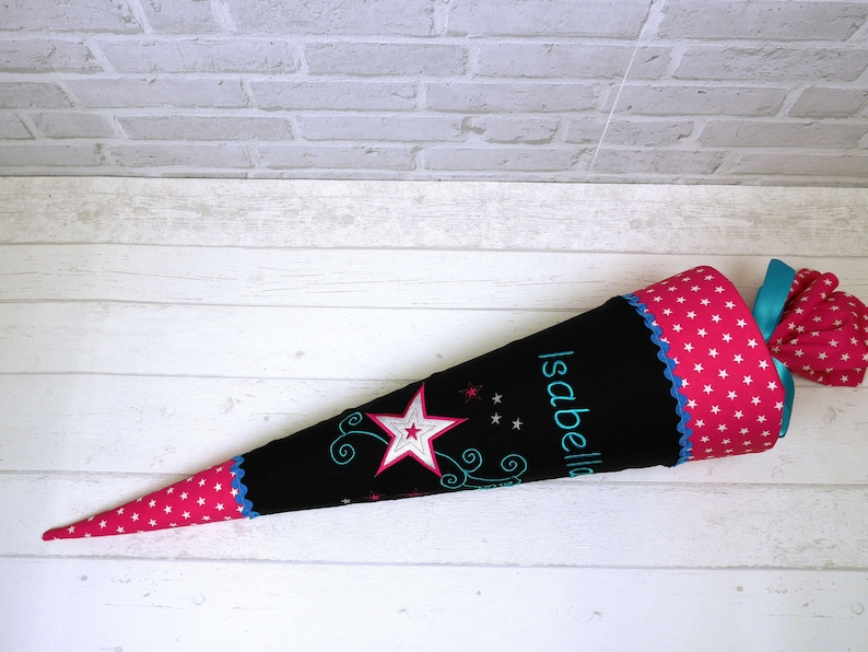 May include: A black and pink school cone with white stars and the name "Isabella" embroidered on it. The cone has a pink and white polka dot pattern and a blue ribbon.