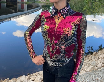 May include: A black cowboy hat tops a long-sleeved, sequined shirt in shades of red, pink, and gold, with a black collar and cuffs. The shirt is paired with black pants and a silver belt. The background features a bridge and a body of water.