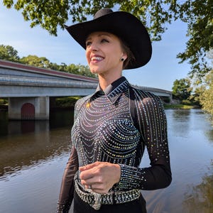 May include: A woman wearing a black cowboy hat, a black long-sleeved shirt with rhinestones, and black pants with a rhinestone belt. She is standing by a river with a bridge in the background.