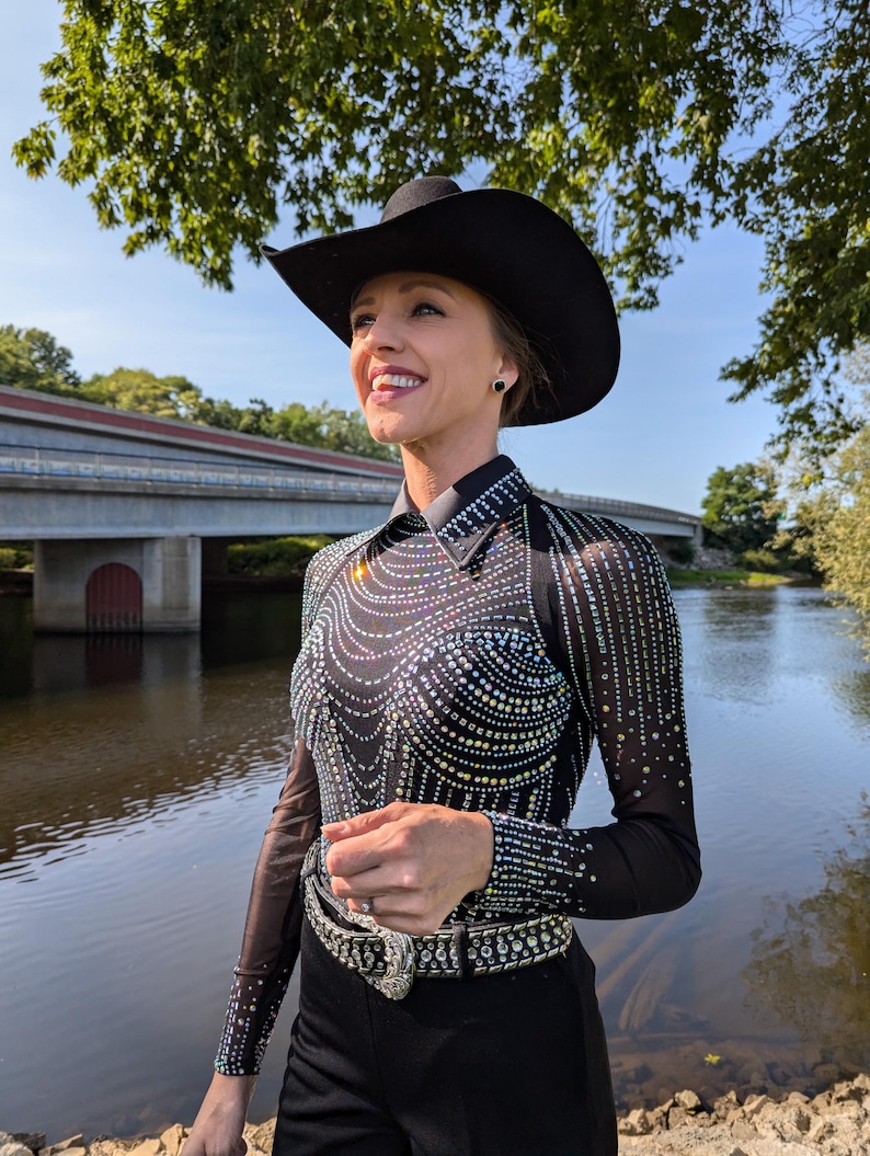 May include: A woman wearing a black cowboy hat, a black long-sleeved shirt with rhinestones, and black pants with a rhinestone belt. She is standing by a river with a bridge in the background.