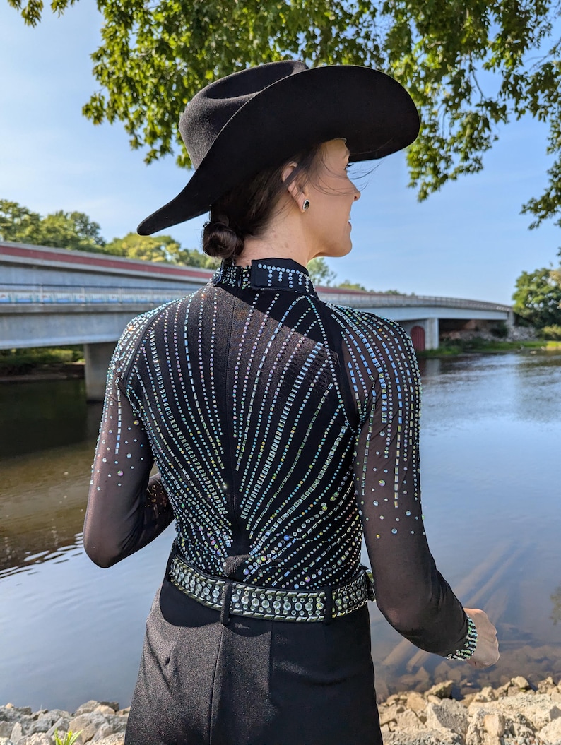 May include: A black long-sleeved shirt with a sequined design and a black belt. The shirt is worn by a person wearing a black cowboy hat. The person is standing in front of a body of water.