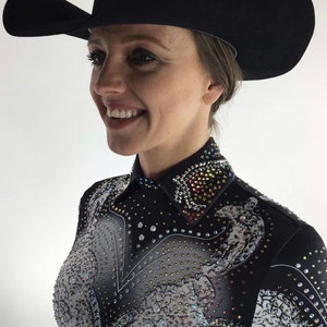 May include: A woman wearing a black cowboy hat with a wide brim and a black and white patterned shirt with rhinestones. The shirt has a pointed collar and a design of a horse on the front.