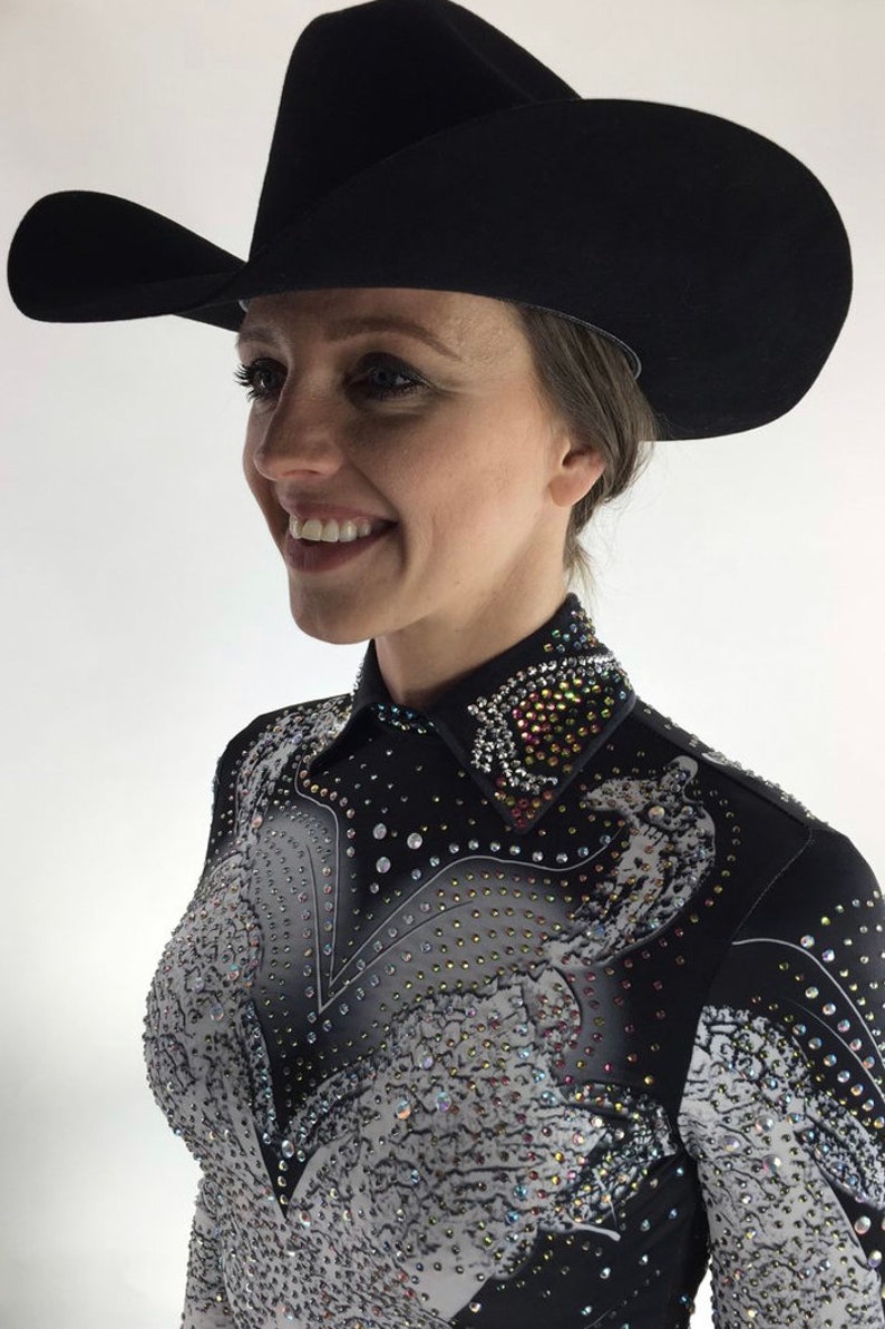 May include: A woman wearing a black cowboy hat with a wide brim and a black and white patterned shirt with rhinestones. The shirt has a pointed collar and a design of a horse on the front.