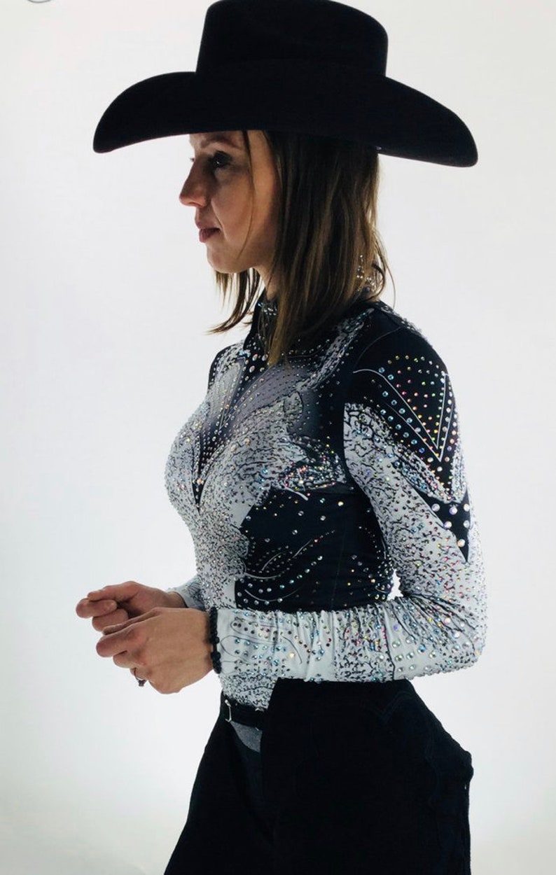 May include: A woman wearing a white and black long-sleeved shirt with rhinestones, black pants, and a black cowboy hat.