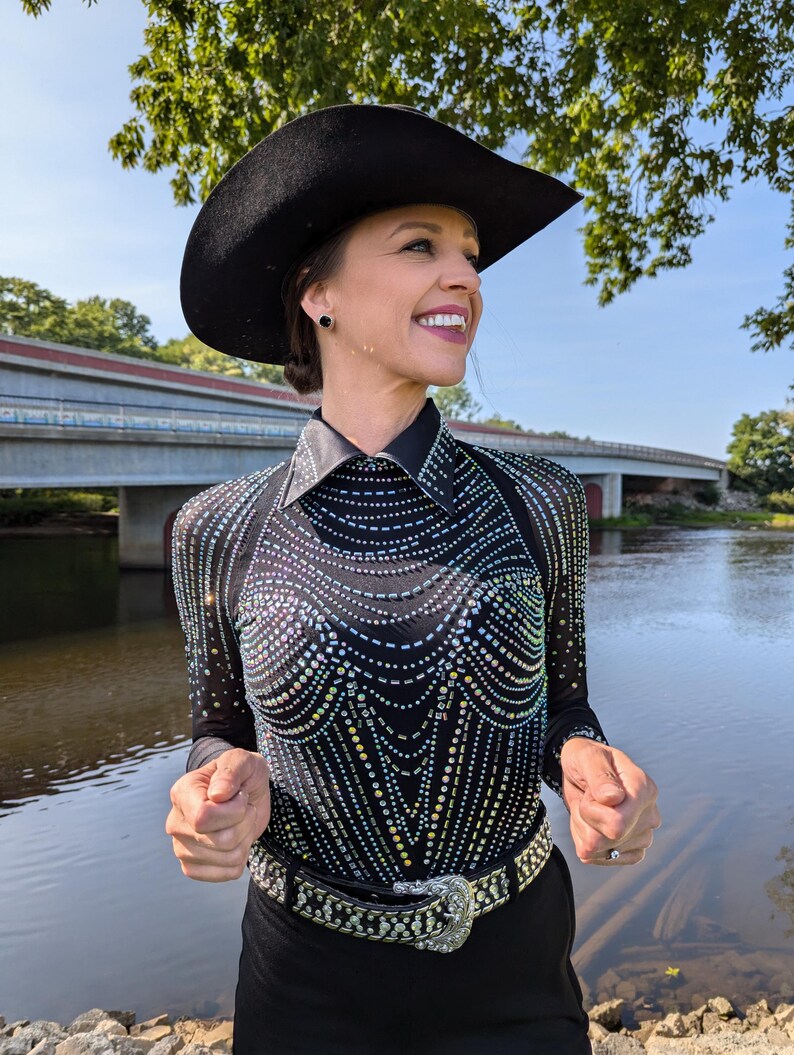 May include: A woman wearing a black cowboy hat, a black sequined top, and a black belt with a silver buckle. She is standing in front of a body of water.