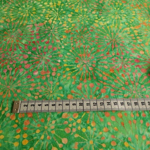 Könnte beinhalten: Grüner Stoff mit einem sich wiederholenden Muster aus orangefarbenen und rosafarbenen stilisierten Feuerwerkskörpern. Ein Maßband liegt auf dem Stoff und zeigt Maße in Zoll.