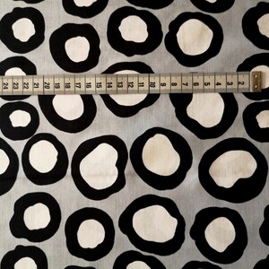 Könnte beinhalten: Grauer Stoff mit einem sich wiederholenden Muster aus schwarzen, umrandeten weißen Kreisen. Ein Maßband ist über den Stoff gelegt und zeigt Maße von 2 bis 28 Zoll.