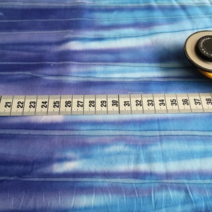 Könnte beinhalten: Nahaufnahme eines blau-weiß gestreiften Stoffes mit einem gelben Maßband und einem Rollschneider. Das Maßband zeigt die Zahlen von 18 bis 41 Zoll.