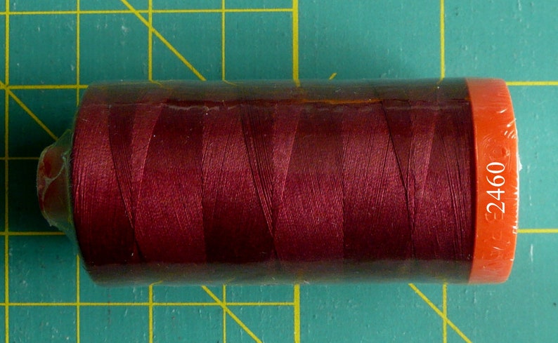 K&ouml;nnte beinhalten: Eine Spule mit maroonfarbenem Garn mit einem wei&szlig;en Etikett, auf dem "2460" auf orangefarbenem Hintergrund steht.