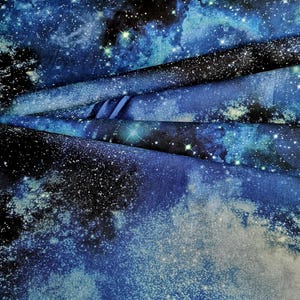 Könnte beinhalten: Ein Stoff mit einem Weltraum-Galaxie-Druck in Marineblau, Königsblau und Schwarz. Das Muster zeigt verstreute weiße Sterne, helle Lichtcluster und nebelartige Wolken, die einen geschichteten Effekt auf dem gefalteten Textil erzeugen.