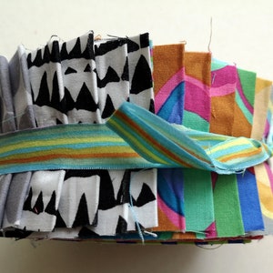 Könnte beinhalten: Ein Stapel bunter Stoffquadrate mit einem geometrischen Muster in Schwarz und Weiß, zusammengebunden mit einem gestreiften Band.