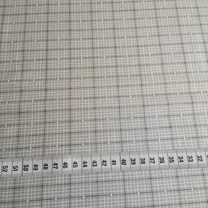 Könnte beinhalten: Ein grauer Stoff mit einem Rastermuster aus weißen Linien und Punkten. Ein Maßband mit Zentimetermarkierungen von 32 bis 52 liegt am Rand des Stoffes. Der Stoff eignet sich für Quilt- oder Nähprojekte.