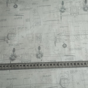 Könnte beinhalten: Grauer und weißer Stoff mit einem sich wiederholenden Muster aus Flugzeugplänen. Der Stoff wird mit einem Maßband gemessen, das Zoll von 1 bis 32 anzeigt.