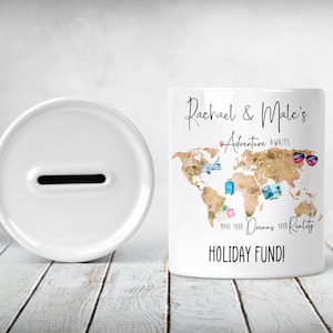 May include: White ceramic money box with a slot on the top. The front of the box features a gold world map with travel icons and the text "Rachael & Marc's Adventure Awaits Make Your Dreams Your Reality HOLIDAY FUND!"