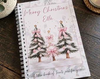 May include: A spiral-bound notebook featuring a white cover with three illustrated Christmas trees decorated with pink bows and ornaments. The text reads Merry Christmas Ella and A little book of Treats just for you! in black and pink script.