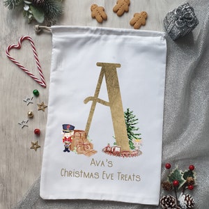 May include: White canvas Christmas Eve treat bag featuring a gold letter A, a nutcracker, presents, and a mouse. The bag is personalised with the name "Ava's" and the text "Christmas Eve Treats".