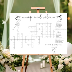 May include: A white sign with the text "Sip and Solve" and a crossword puzzle. The crossword puzzle has clues related to a wedding. The sign also includes the names "Suzie & Daniel" and the date "28.10.25".